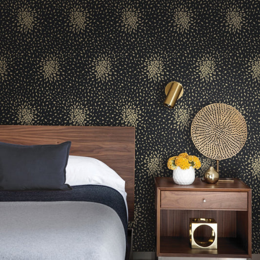 A modern bedroom features York Wallcoverings' Black & Gold Petite Leaves Wallpaper on a feature wall, paired with a wooden headboard and gray-white bedding. A bedside table displays a yellow flower vase, while a metallic lamp and round woven wall decor enhance the contemporary style.