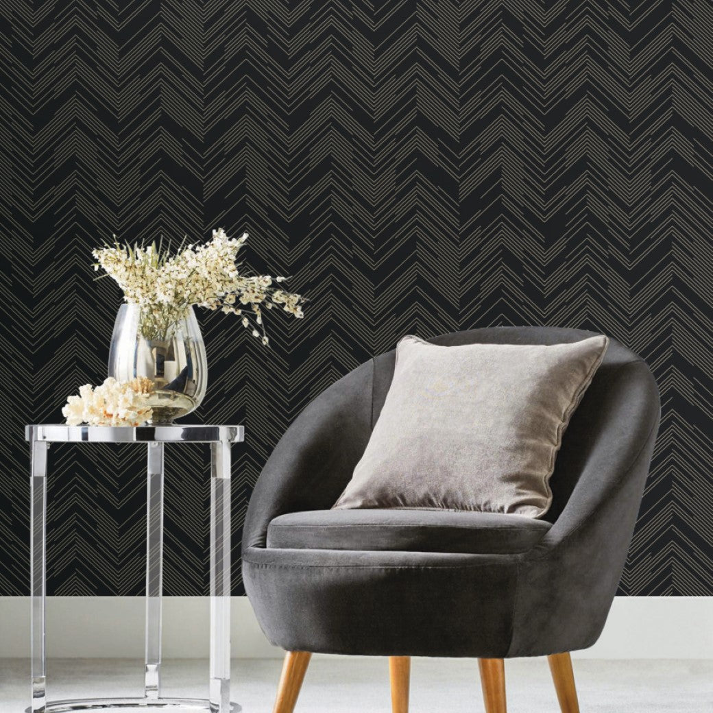 Living Room - A contemporary features a gray upholstered chair with a light gray cushion beside a transparent round side table holding a glass vase of white flowers. Metallic York Wallcoverings' Black & Gold Polished Chevron Wallpaper adorns the background, adding sleek sophistication