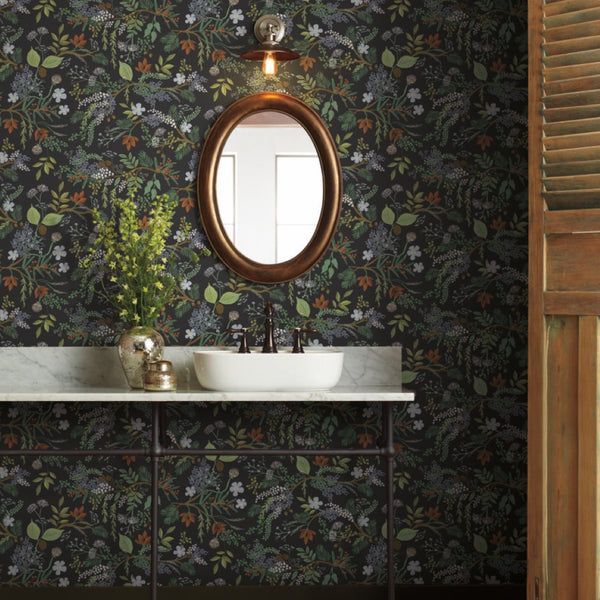 Bathroom - A features a white sink with a dark faucet on marble, a vase with greenery, and a round gold-framed mirror beneath a light, set against York Wallcoverings’ Black Juniper Forest Wallpaper for a stunning nature-inspired touch