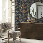 Living Room - A cozy showcases a beige armchair with a chunky knit throw, a wooden sideboard, a round mirror, and an ornamental plant. The backdrop features York Wallcoverings' Black & Multi Shimmering Foliage Wallpaper, adding depth and elegance. A spiral staircase is in the background