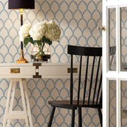 A white desk with gold accents holds a lamp and a vase of white flowers. A black chair sits in front, backed by York Wallcoverings' Blue/White Laurel Wallpaper (60 SqFt) and a glass-paned door partially visible.