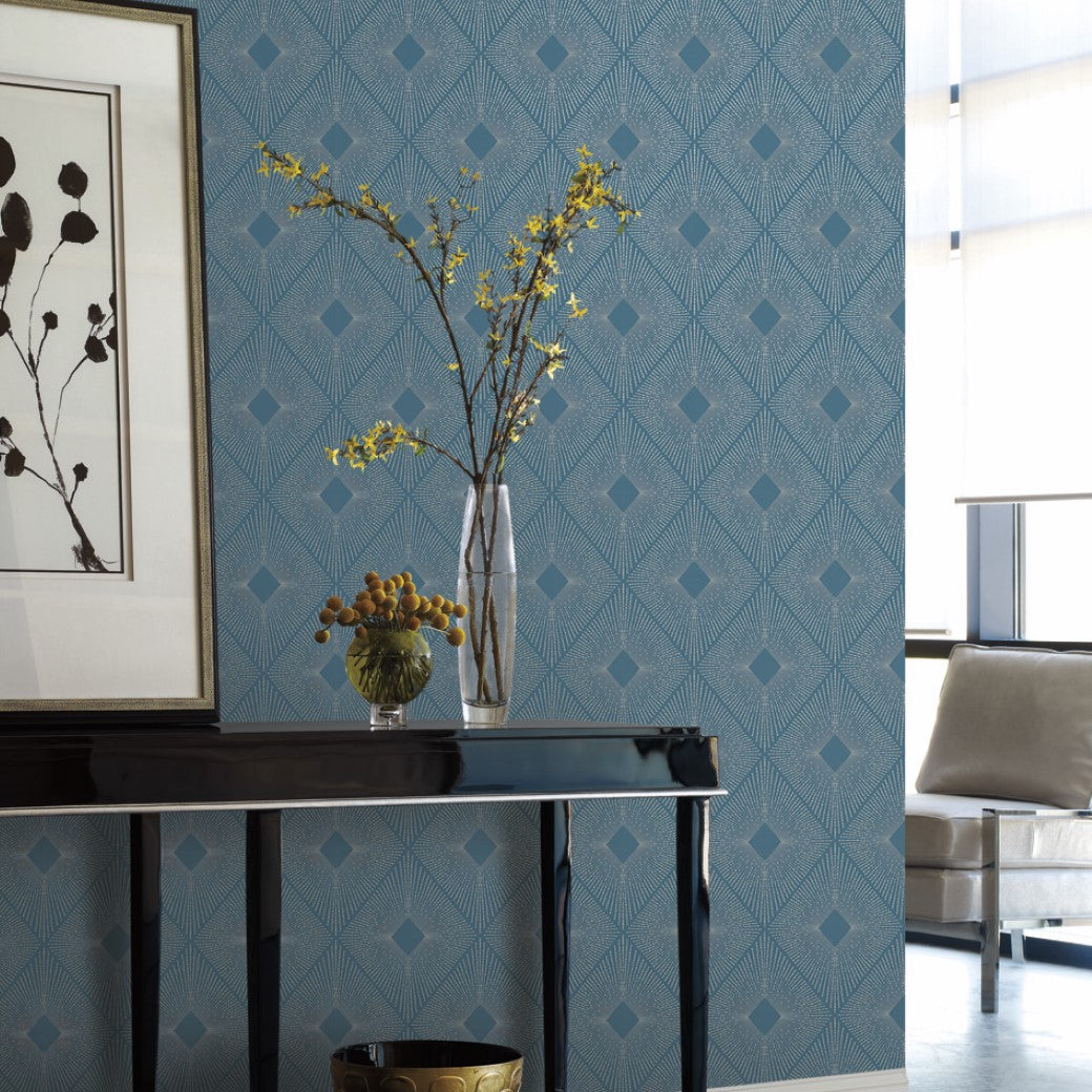 A modern space featuring York Wallcoverings Blue & Silver Harlowe Wallpaper showcases framed botanicals. A black console table adds metallic elegance with a tall vase of yellow blooms and a small grape bowl. Large windows bathe the room in natural light.