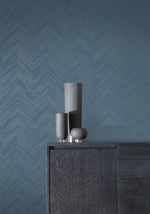 Three sleek dark vases of different heights sit on a dark wooden cabinet against York Wallcoverings' Blue & Silver Polished Chevron Wallpaper, adding metallic accents that enhance the minimalist modern aesthetic of this space.