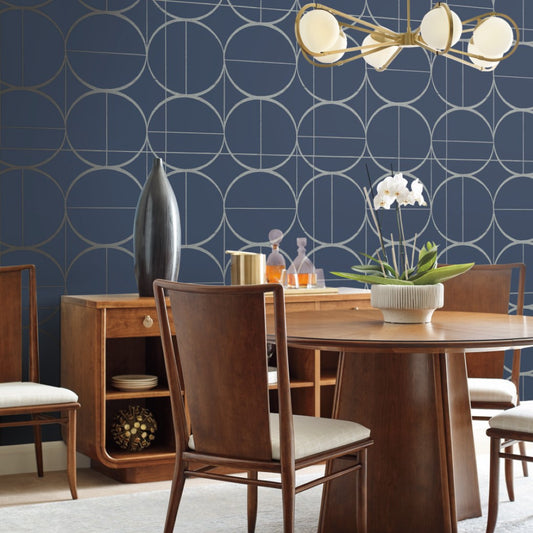 A modern dining room includes a round wooden table with four chairs and metallic accents for sophistication. A sideboard holds decor and a vase, while the York Wallcoverings' Blue & Silver Sun Circles Wallpaper highlights the gold chandelier. The table features a white orchid centerpiece.