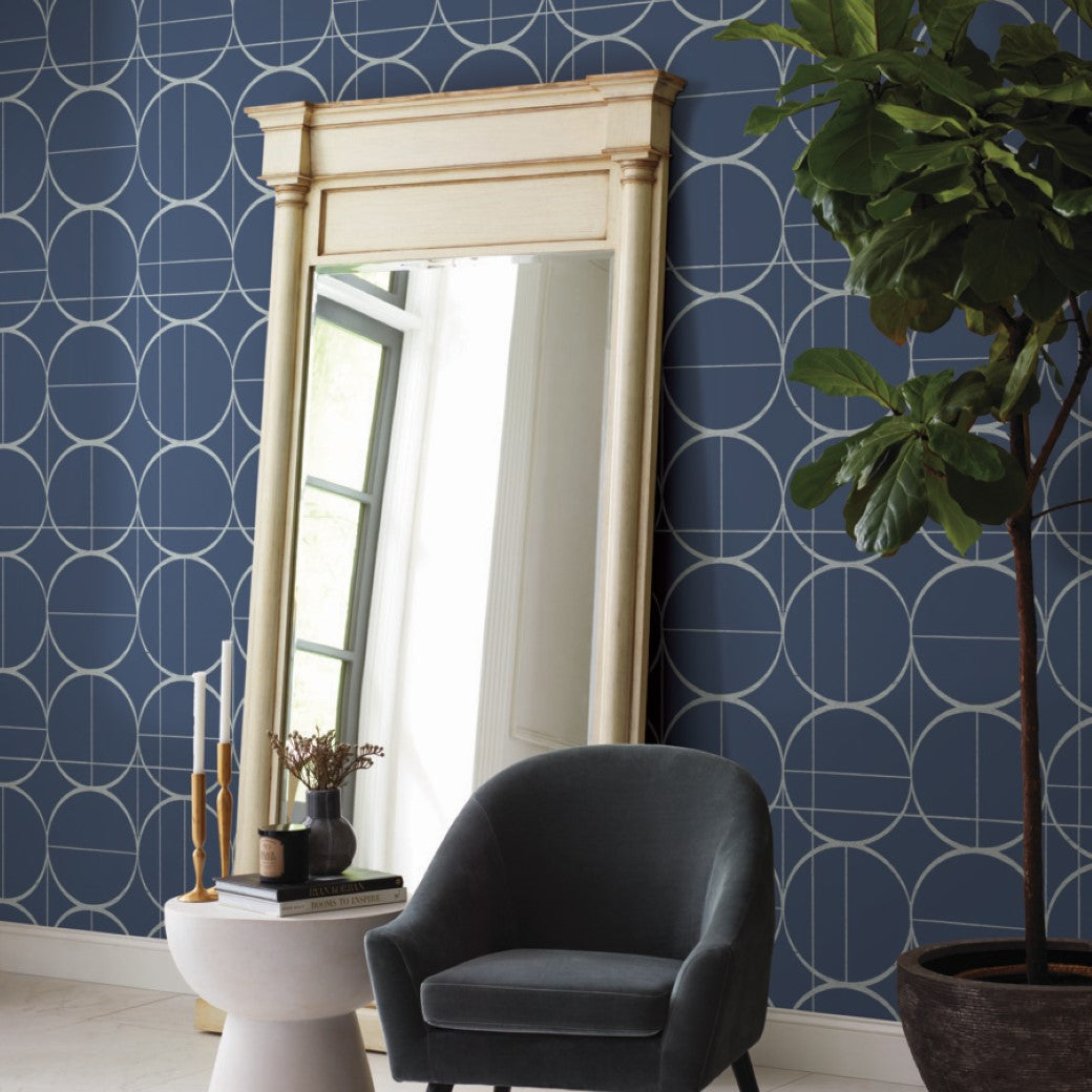 A tall wooden-framed mirror leans against a wall with York Wallcoverings' Blue & Silver Sun Circles wallpaper. Beside a small round white table with books, candles, and a plant sits a dark gray armchair. A lamp glints with metallic finishes, and a tall potted plant stands in the corner.