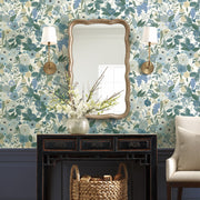 A room with Blues Garden Party Wallpaper by York Wallcoverings features a scalloped mirror, wall sconces, a dark wood console with white flowers and a wicker basket below, plus a beige armchair on the right.