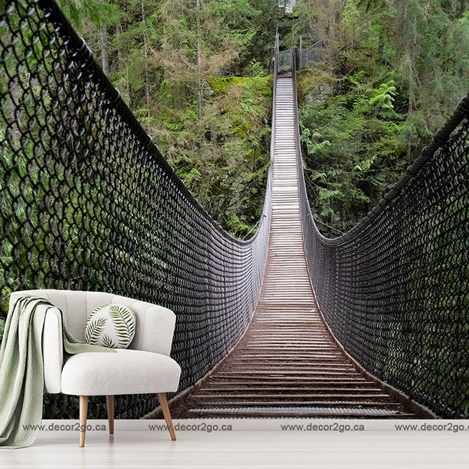 A surreal indoor scene blending a Bridge Amid the Forest Wallpaper Mural from Decor2Go Wallpaper Mural with a suspension bridge and a cozy home setting, featuring a stylish chair, a throw blanket, a cushion, and a small table with a plant.