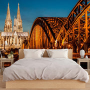 Bedroom - A creative bedroom setup with a wall-sized Bridge and Cathedral View Wallpaper Mural from Decor2Go Wallpaper Mural depicting the cologne cathedral and hohenzollern bridge at night, featuring a bed with white bedding in the foreground, designed to evoke.