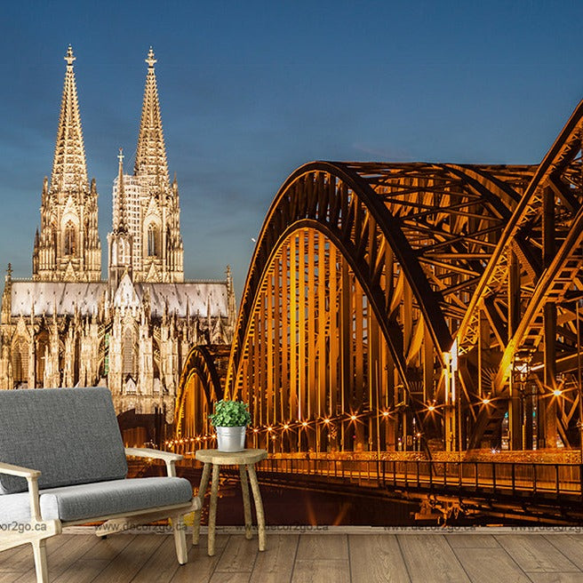 A surreal composition featuring a modern armchair and side table with a plant positioned on a wooden floor, seamlessly blended in front of the illuminated Bridge and Cathedral View Wallpaper Mural at dusk under a Decor2Go Wallpaper Mural.