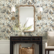 A room with York Wallcoverings’ Brown/Beige Garden Party Wallpaper features a black console table, scalloped mirror, wall sconces with white shades, a white vase of flowers, woven basket below, and part of a beige chair on the right.
