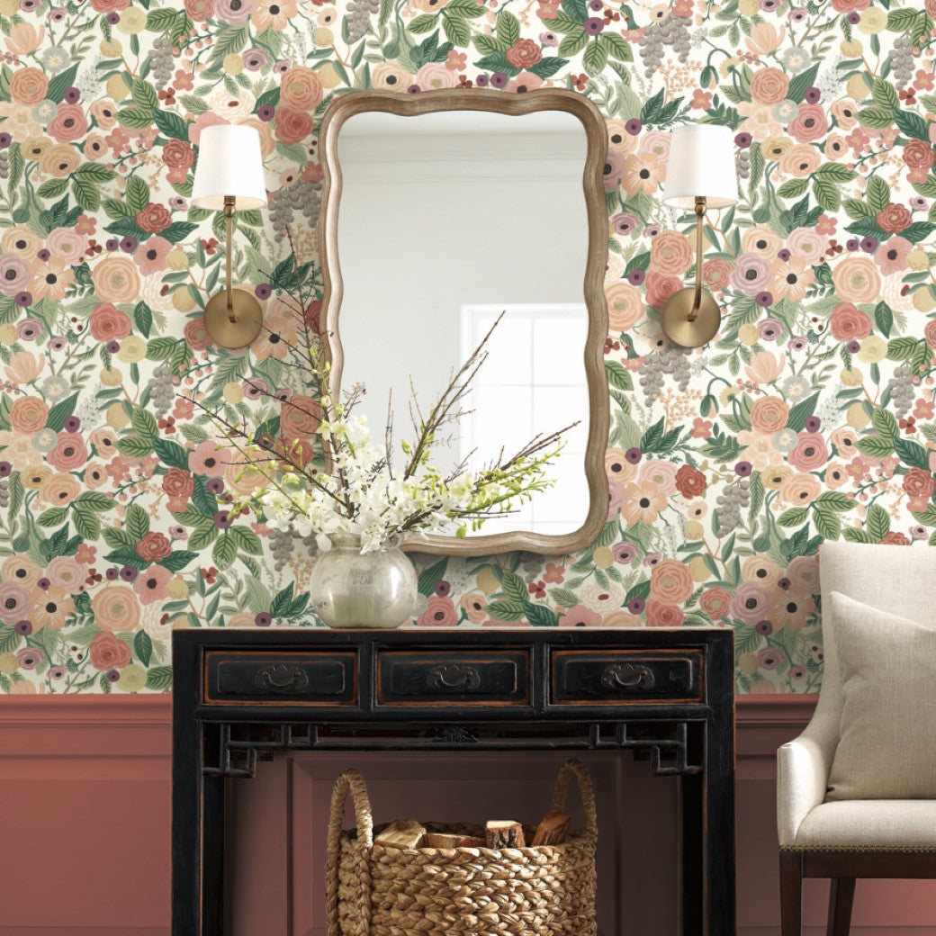 A mirror with a wavy wood frame is mounted on York's Burgundy Garden Party Wallpaper, showcasing botanical artistry. Two wall sconces flank it. Below, a chest of drawers supports a vase of flowers. A wicker basket sits at the base, with part of a beige couch visible in the corner.