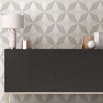 A modern, black sideboard stands against a wall with York Wallcoverings' Contemporary Silver and Grey Geometric Wallpaper (56 SqFt) featuring a geometric, grey and white pattern. On top of the sideboard, there is a sleek white and grey marble lamp, several decorative books, and a small grey spherical ornament, embodying modern elegance.