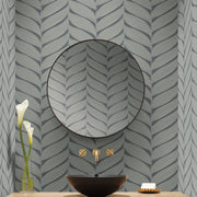 Bathroom - A modern features a large round mirror, an angled gold faucet, and a black bowl-shaped sink. Two calla lilies are in a vase on the left, with a soap dish on the right. The wall showcases York Wallcoverings' Charcoal & Silver Luminous Leaves Wallpaper