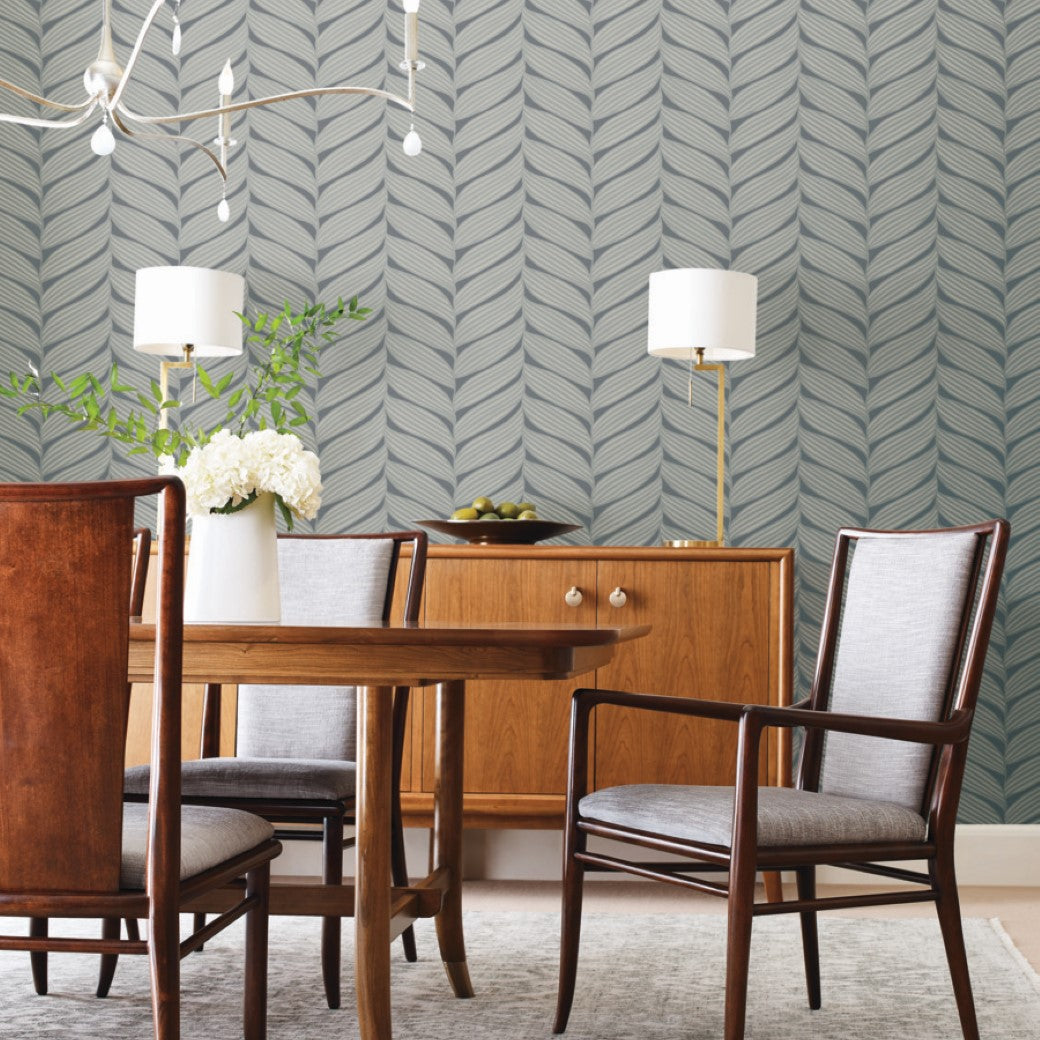 A dining room with a wooden table and four chairs showcases metallic details. Against York Wallcoverings' Charcoal & Silver Luminous Leaves Wallpaper sits a sideboard with lamps and apples. Overhead, a modern light fixture casts a glow on a vase of white flowers.