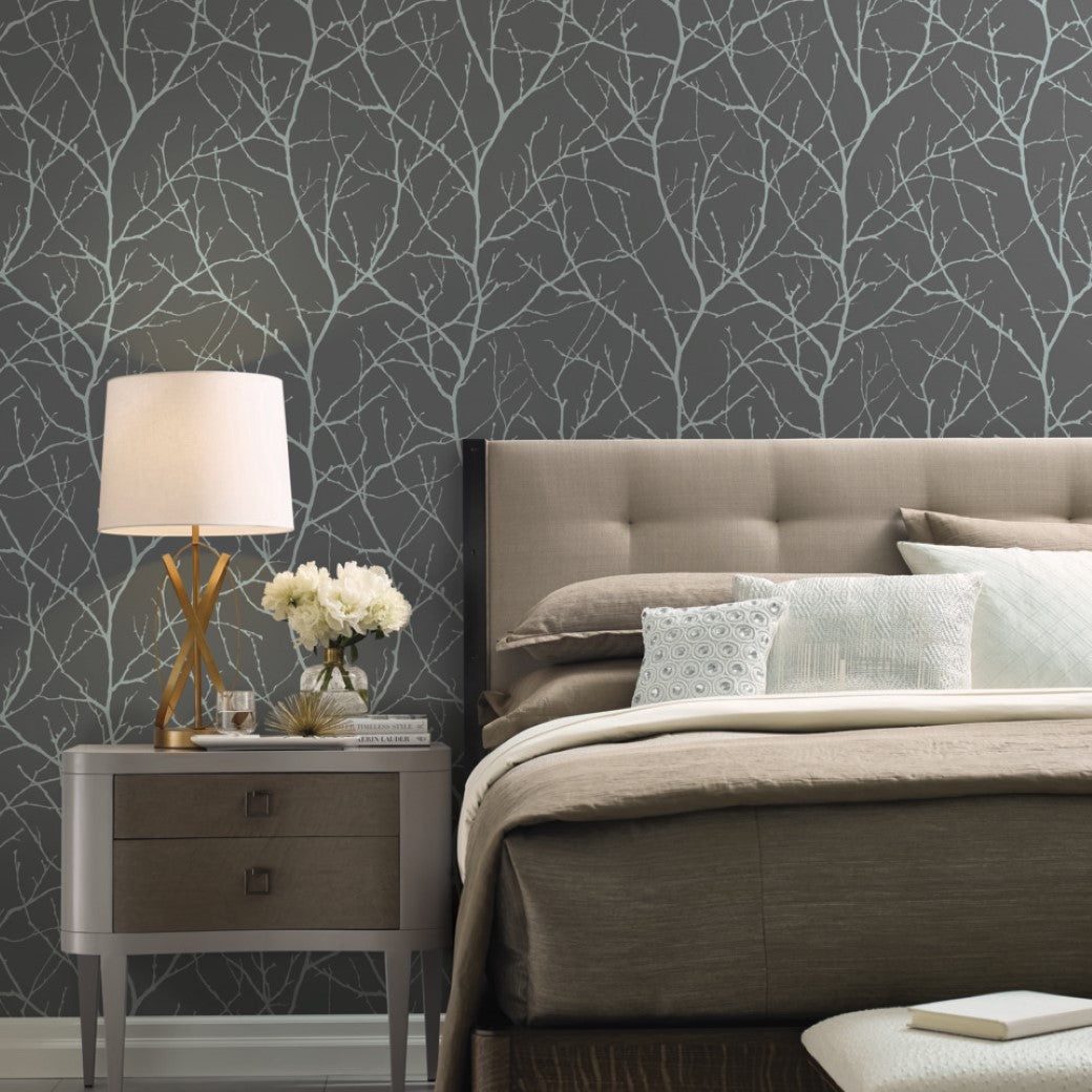 A modern bedroom features York Wallcoverings' Charcoal & Silver Trees Silhouette Wallpaper, paired with a beige bed, white and light blue pillows, and a wooden nightstand holding a white lamp and vase of white flowers, perfectly capturing the sophistication of the Modern Metals collection.