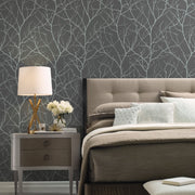 Bedroom - A modern features York Wallcoverings' Charcoal & Silver Trees Silhouette Wallpaper, paired with a beige bed, white and light blue pillows, and a wooden nightstand holding a white lamp and vase of white flowers, perfectly capturing the sophistication of the Modern Metals collection