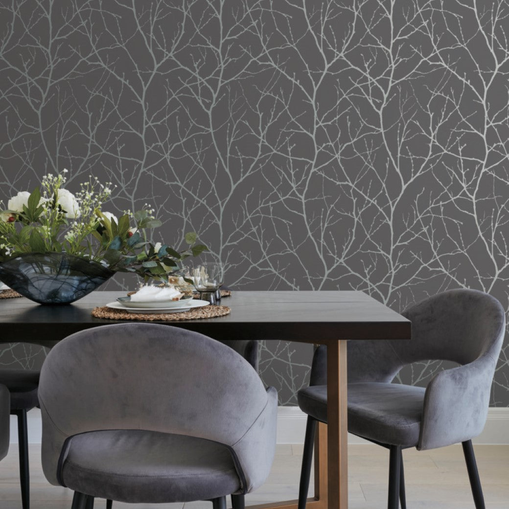 A modern dining area features a dark wood table with gray upholstered chairs. The backdrop is Charcoal & Silver Trees Silhouette Wallpaper by York Wallcoverings, featuring a Tree Silhouette pattern. A decorative bowl with greenery adorns the table.