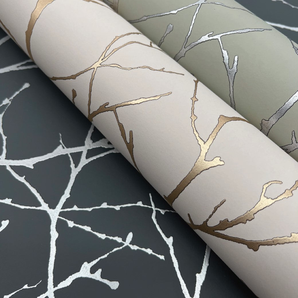 Two rolls from York Wallcoverings' Modern Metals collection are displayed on a dark surface, showcasing metallic tree branch designs: one in gold on beige, and the Charcoal & Silver Trees Silhouette design on light green.