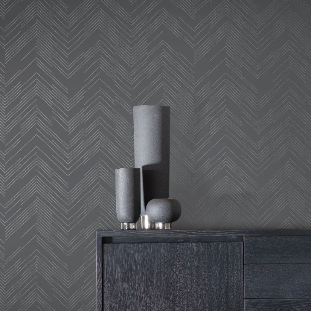 A set of three dark gray ceramic vases of varying heights sits atop a dark wooden cabinet. Behind them, York Wallcoverings' Charcoal & Silver Polished Chevron Wallpaper adds a modern, minimalist aesthetic.