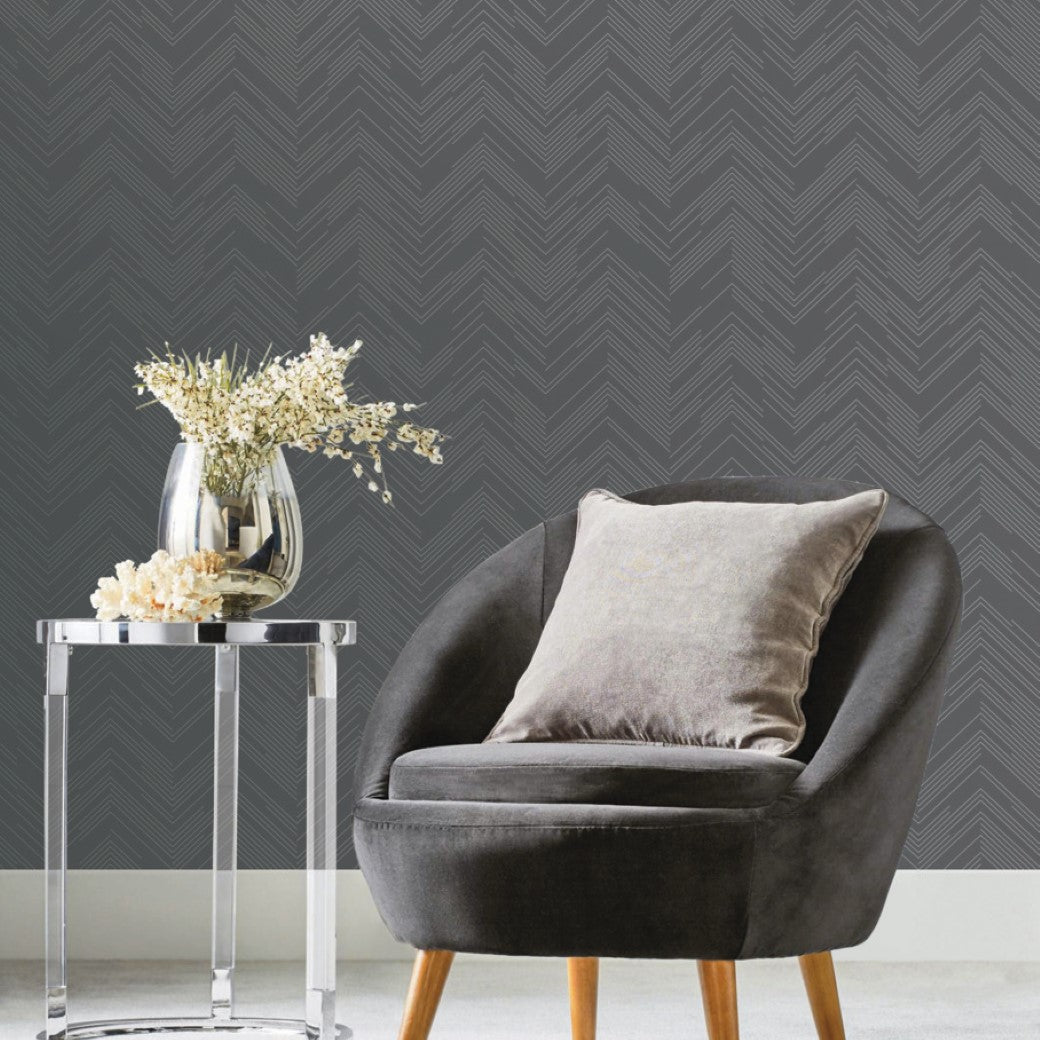 A modern room with a gray, cushioned armchair and a gray pillow next to a clear glass side table supporting a vase of white flowers. The wall features York Wallcoverings' Charcoal & Silver Polished Chevron Wallpaper, enhancing the minimalist decor.