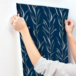 Person wearing a striped shirt, seen from the back, aligning a dark blue York Wallcoverings Chloe Vine Indigo Wallpaper Blue, Green (60 Sq.Ft.) with a vertical leaf pattern on a white wall. The whimsical design requires careful hands to hold the top corners of the SureStrip wallpaper for proper placement.