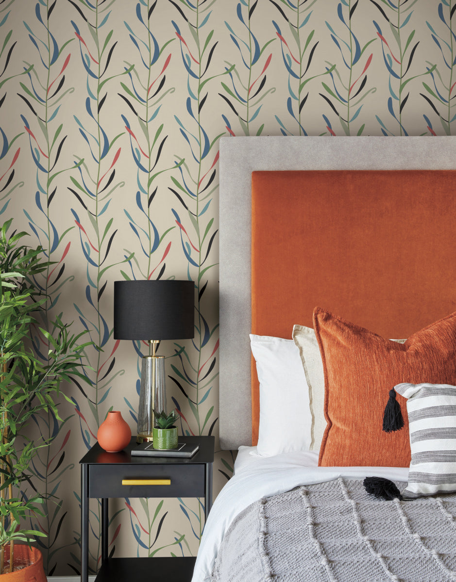 A cozy bedroom features a bed with an orange upholstered headboard and matching pillows. There is a black nightstand with a white vase, a glass lamp with a black shade, and a small orange sculpture. The York Wallcoverings Chloe Vine Indigo Wallpaper Blue, Green (60 Sq.Ft.) has a leafy pattern, adding to the whimsical design alongside the potted plant beside the bed.