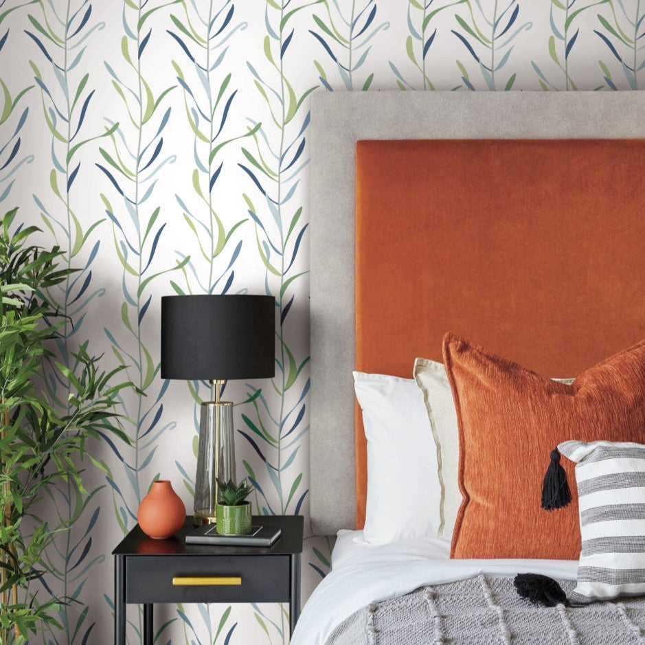 A cozy bedroom featuring a bed with an orange headboard and various pillows in shades of orange, white, and gray. A black nightstand with a black lamp, a small terracotta vase, and potted greenery sits beside the bed. The York Wallcoverings Chloe Vine Indigo Wallpaper Blue, Green (60 Sq.Ft.) adds a whimsical design to the leafy-patterned walls.