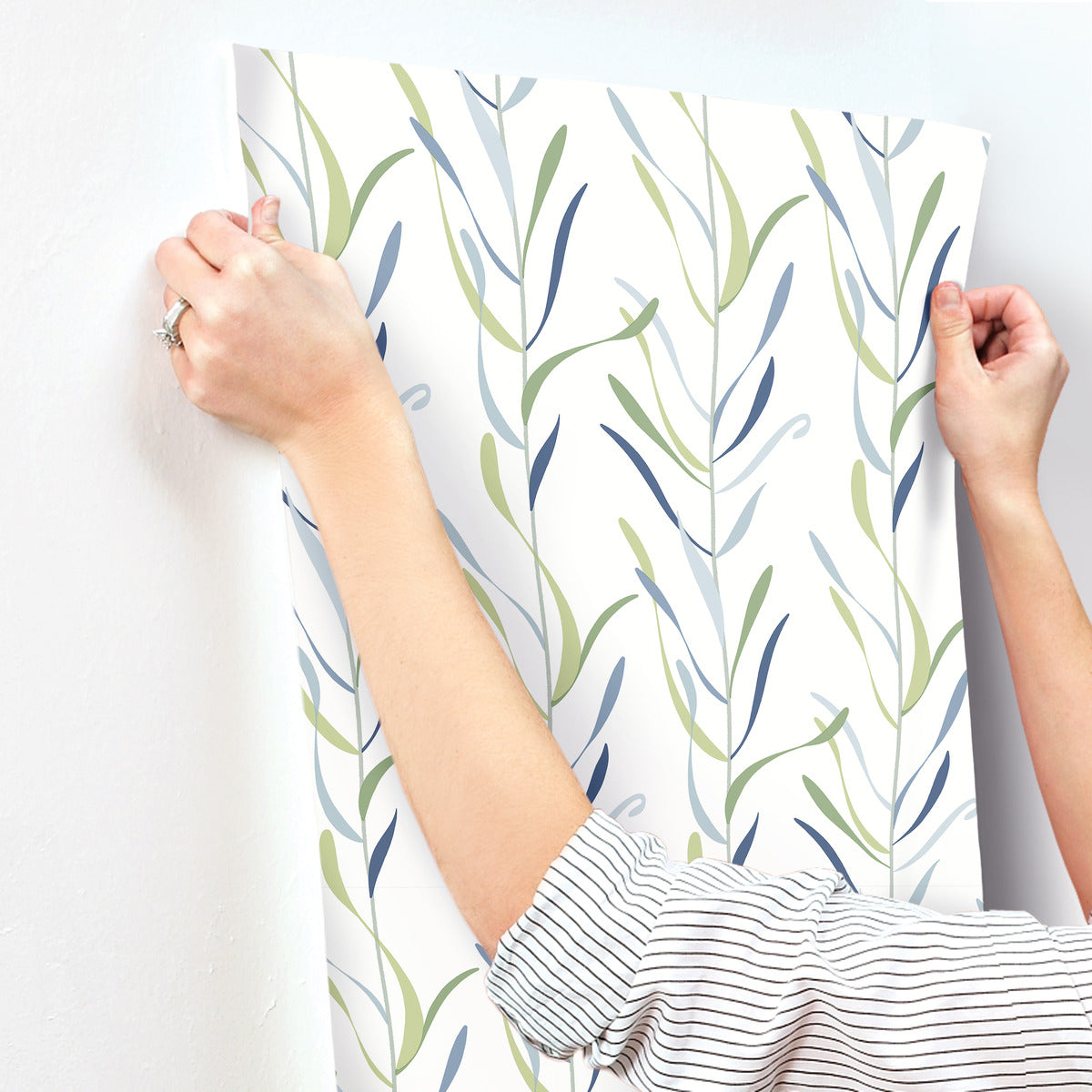 A person wearing a striped shirt is applying York Wallcoverings Chloe Vine Indigo Wallpaper Blue, Green (60 Sq.Ft.) with a whimsical design of green and blue leaves to a white wall. The person's hands are smoothing out the wallpaper, and a ring is visible on their finger.