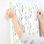 A person hanging a large sheet of Chloe Vine River Rock Wallpaper Beige, Grey (60 Sq.Ft.) with a green and blue leaf pattern by York Wallcoverings on a white wall.