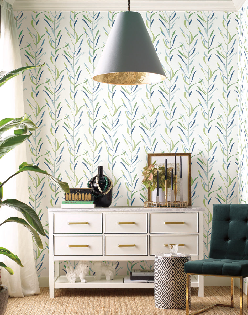 A stylish living room features a white sideboard with gold handles, holding decorative items including a vase, greenery, books, and candles. Above, a large pendant light hangs. A green upholstered chair and a patterned stool are nearby. The wall is adorned with Chloe Vine Indigo Wallpaper Blue, Green (60 Sq.Ft.) by York Wallcoverings in a whimsical design.