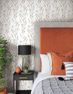 A modern bedroom with York Wallcoverings' Chloe Vine Indigo Wallpaper Blue, Green (60 Sq.Ft.) featuring a leafy-patterned whimsical design, an orange upholstered headboard, and neutral bedding. The bedside table showcases a black lamp with a white shade, a potted plant, and a small orange vase. A green plant sits beside the table.