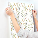 A person with long sleeves installs Chloe Vine Indigo Wallpaper Blue, Green (60 Sq.Ft.) by York Wallcoverings with a leafy pattern on a white wall. The wallpaper, featuring a whimsical design with green, brown, and grey leaves, is likely SureStrip. The person's ring indicates they are married. The image focuses on their hands and the wallpaper pattern.