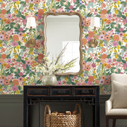A decorative room featuring Coral/Orange Garden Party Wallpaper by York Wallcoverings, a scalloped-edge wall mirror, two brass sconces, a black console table with white flowers and basket beneath, plus a beige armchair.