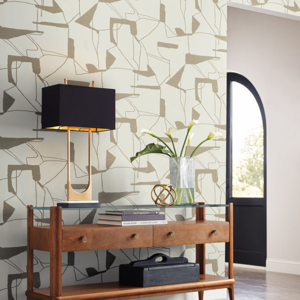 A minimalist interior scene showcases a wooden console table with two drawers. Atop, a black lamp with a unique base sits alongside a vase of green leaves and a decorative object. The wall features Warm Grey & Gold Harlowe Wallpaper by York Wallcoverings, complemented by an arched window.