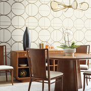 Dining Room - A modern features a round wooden table, four chairs, a large vase, and a potted orchid. The wall displays York Wallcoverings' Cream & Gold Sun Circles Wallpaper in metallic gold circles, complemented by a contemporary chandelier above the table