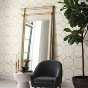 A modern interior features a large ornate mirror against a York Wallcoverings Cream & Gold Sprigs Wallpaper wall. In front sits a small white table with decor, alongside a dark gray chair and a tall green plant to the right.