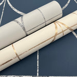 Two rolls of York Wallcoverings' Cream & Gold Sun Circles Wallpaper feature exquisite geometric designs. One roll has silver on cream, the other metallic gold on beige. Both rest elegantly on a dark blue surface with white geometric lines.