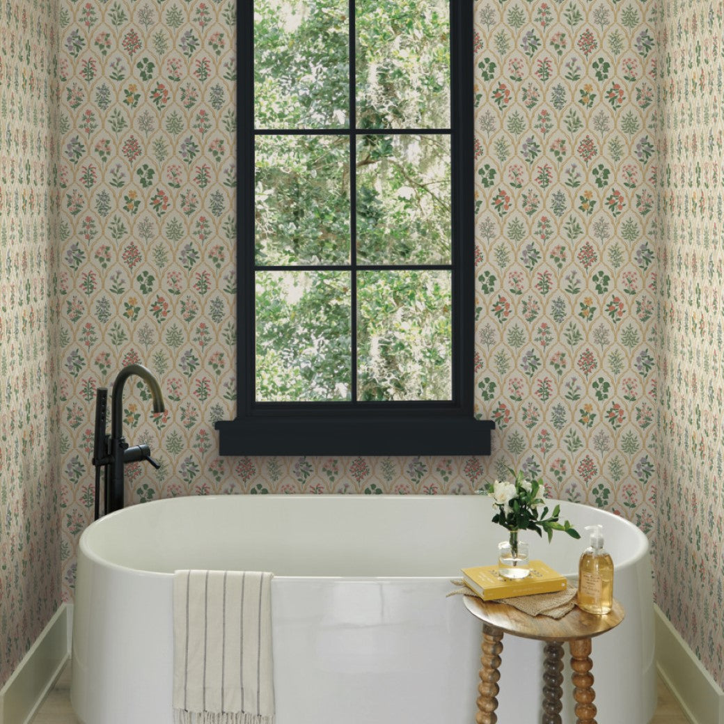 A white freestanding bathtub sits below a large black-framed window with a view of green trees. The wall is adorned with Cream Hawthorne Wallpaper (60 SqFt) by York Wallcoverings. A small wood table holds a plant, soap dispenser, and glass; a towel hangs on the tub.