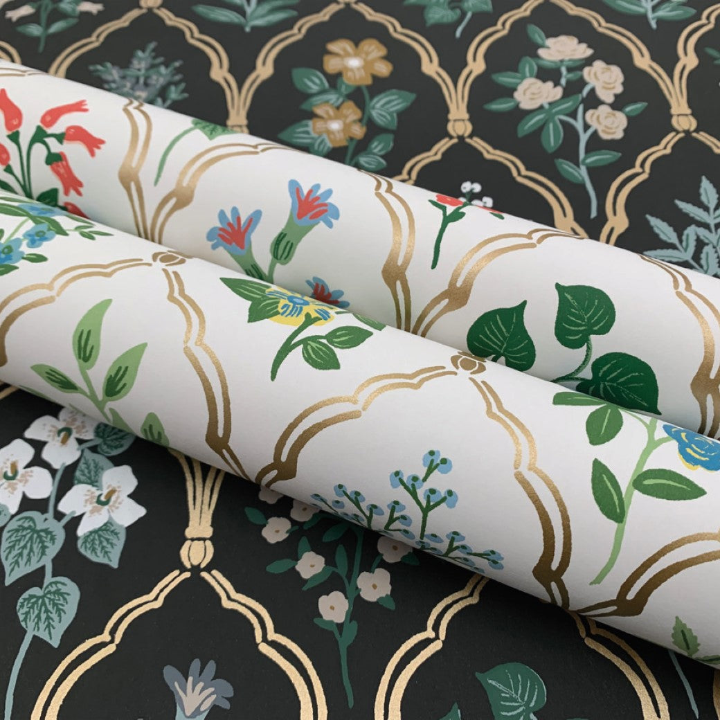 Two rolls of York Wallcoverings Cream Hawthorne Wallpaper (60 SqFt), featuring gold arches and floral designs, rest on a black surface with a matching vintage pattern adorned with vibrant flowers and lush green leaves.