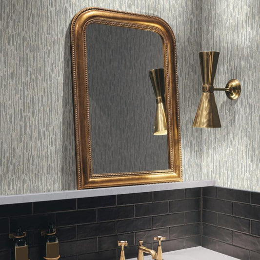 A bathroom interior features a white sink with gold faucets, a rectangular gold-framed mirror, and a gold wall sconce. The York Wallcoverings Cream & Silver Metallic Cascade Wallpaper enhances the textured pattern, while the white stone counter with black tile accents creates a modern high-impact space.