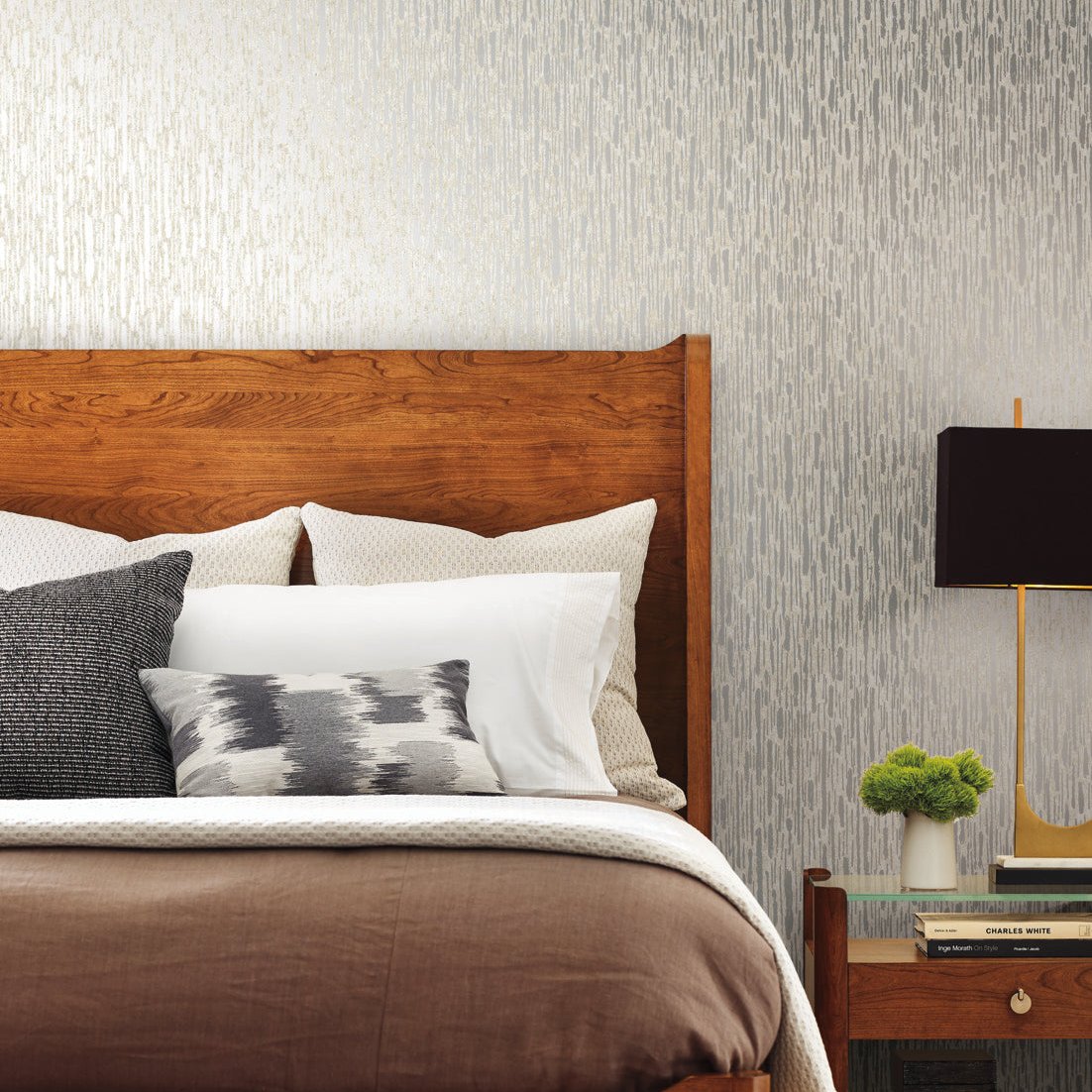 A neatly made bed with a wooden headboard features various pillows and a brown blanket. Beside the bed, a wooden nightstand holds a modern black lamp and small plant. The background showcases York Wallcoverings' Cream & Silver Metallic Cascade Wallpaper, adding elegance to the modern space.