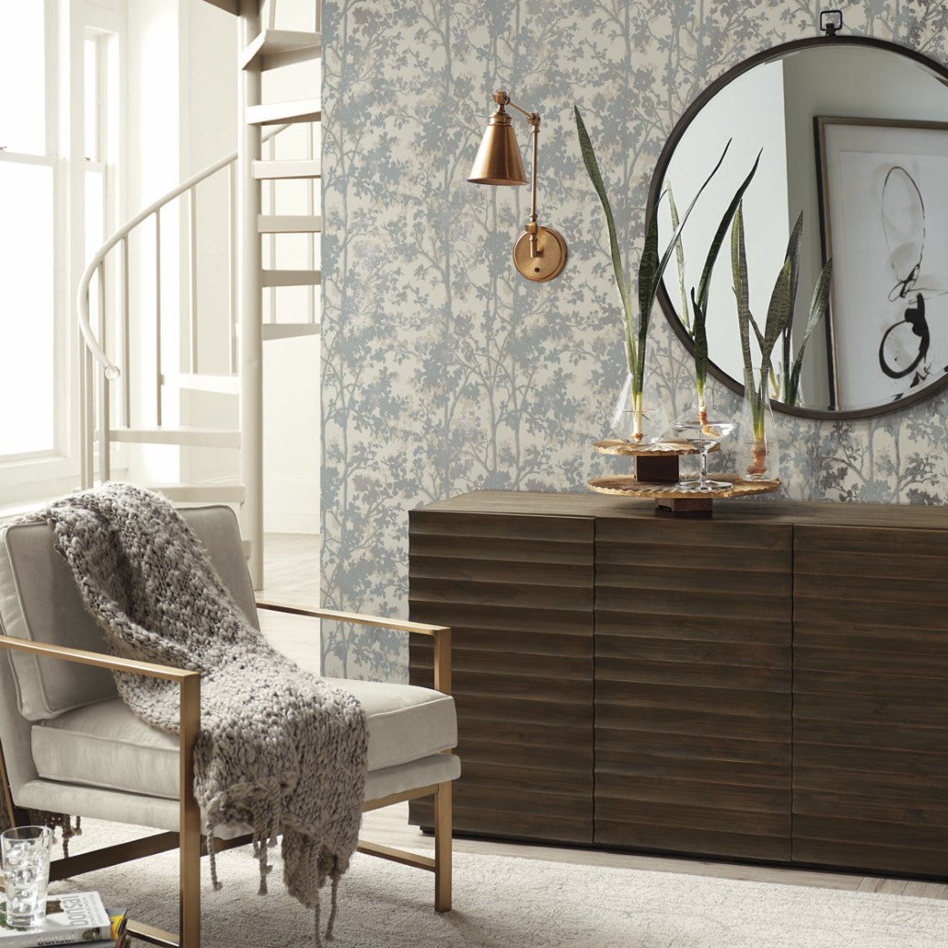 A cozy living room features a wooden sideboard, round mirror, and York Wallcoverings' Cream & Silver Shimmering Foliage Wallpaper. A white armchair with a gray knit throw is on the left, with a Modern Metals Collection copper wall lamp above. A spiral staircase and large windows are in the background.