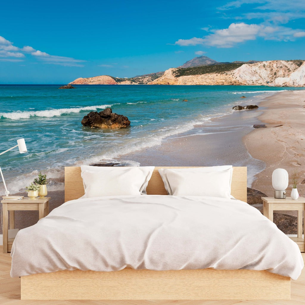 A surreal image of a bed with white bedding on a sandy beach, positioned near the water with a backdrop of mountains and a clear blue sky, evoking a calming atmosphere is captured in the Decor2Go Wallpaper Mural "Crystal Coast Wallpaper Mural.