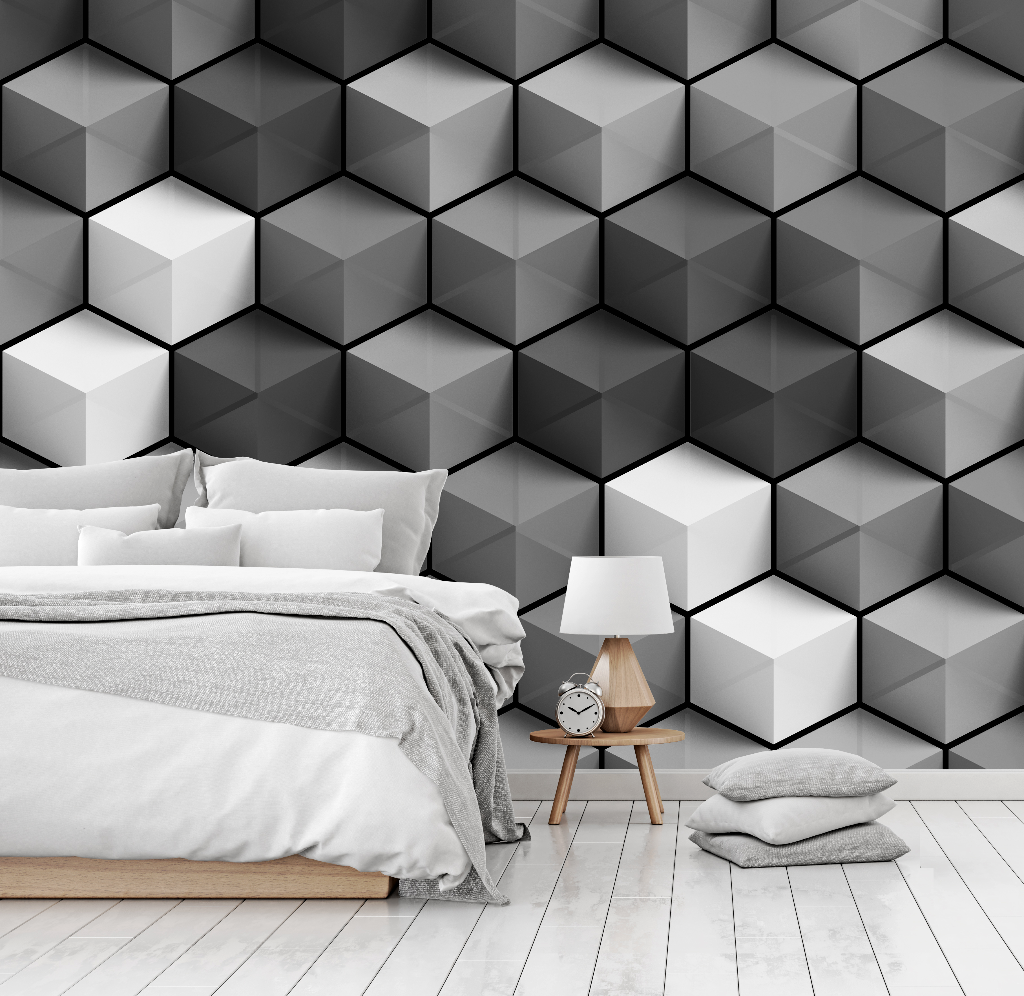 A modern bedroom features a neatly made bed with gray bedding against a Decor2Go Wallpaper Mural featuring Cubic Grays geometric wallpaper. A wooden bedside table with a lamp and clock, and two gray cushions lie on the