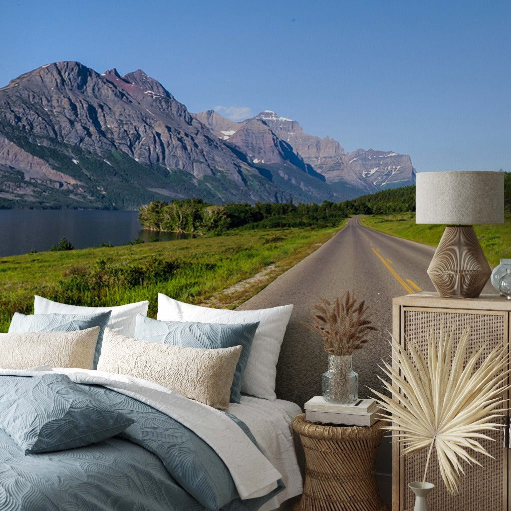 A surreal image combining a scenic mountain road with a cozy bedroom, featuring a bed with blue and beige bedding, a lamp, and decorative items on a wooden nightstand against Decor2Go Wallpaper Mural.