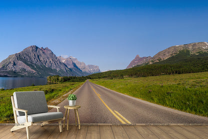 A serene landscape shows an empty road stretching toward distant mountains under a clear blue sky. To the left, a vibrant blue lake reflects the surroundings. In the foreground are a modern armchair and small table, placed on a wooden floor, like a scene from an adventurous Runaway Wallpaper Mural by Decor2Go Wallpaper Mural.