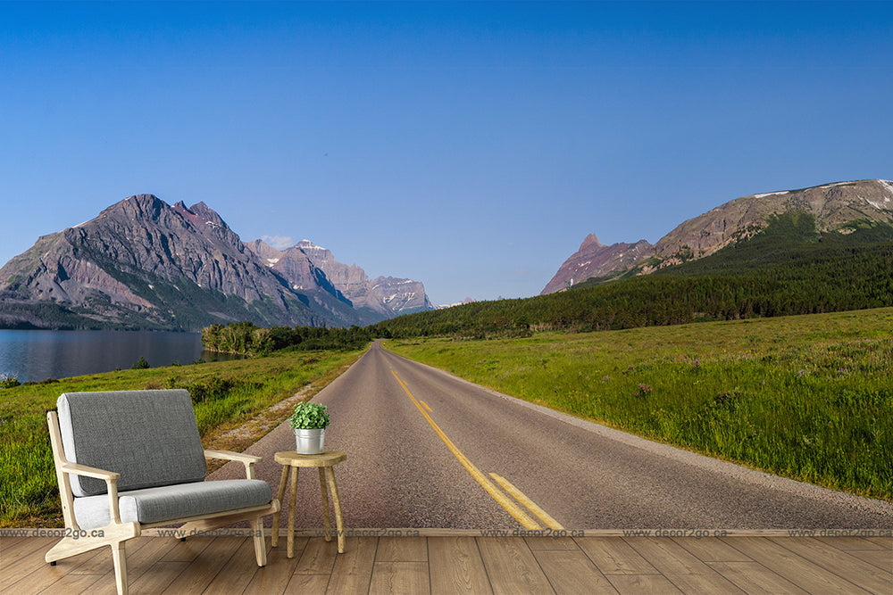 A Decor2Go Wallpaper Mural featuring a scenic mountain landscape beside a lake with a road leading through it. An armchair and a side table with a plant are oddly placed on a rustic wooden platform on the roadside