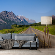 Living Room - A wall mural in a modern living room features an adventurous mountain scape under a clear blue sky. The interior includes a gray couch with a checkered blanket, a small round side table, and a tall floor lamp with a beige shade decorated with the **Runaway Wallpaper Mural by Decor2Go Wallpaper Mural**.