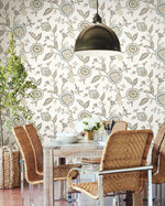 A cozy dining area with a wooden table and four brown wicker chairs with metal frames. The table is set with white dishes, glass jars, and a small bouquet of white flowers. A potted plant stands nearby, and the wall features York Wallcoverings Dahlia Blooms Dove/Cornflower Wallpaper Blue, White/Off White (60 Sq.Ft.) that adds an easy installation touch of botanical elegance.