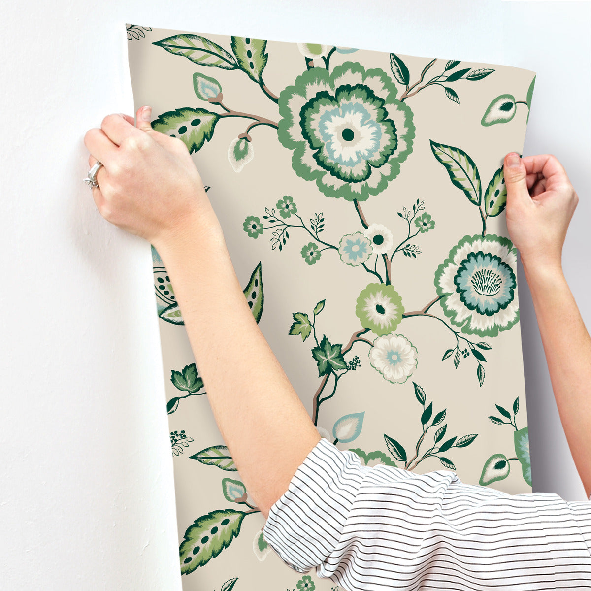 A person with a striped shirt is hanging York Wallcoverings Dahlia Blooms Dove/Cornflower Wallpaper Blue, White/Off White (60 Sq.Ft.), showcasing an intricate floral pattern with green and teal flowers and leaves on a light beige background. The wallpaper exudes botanical elegance as the person's hands carefully hold it at the edges near a white wall.